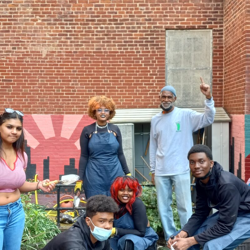 Teacher Earl Grey and four high school students stand in front of a colorful mural on a brick wall posing with their freshly weeded school garden beds.