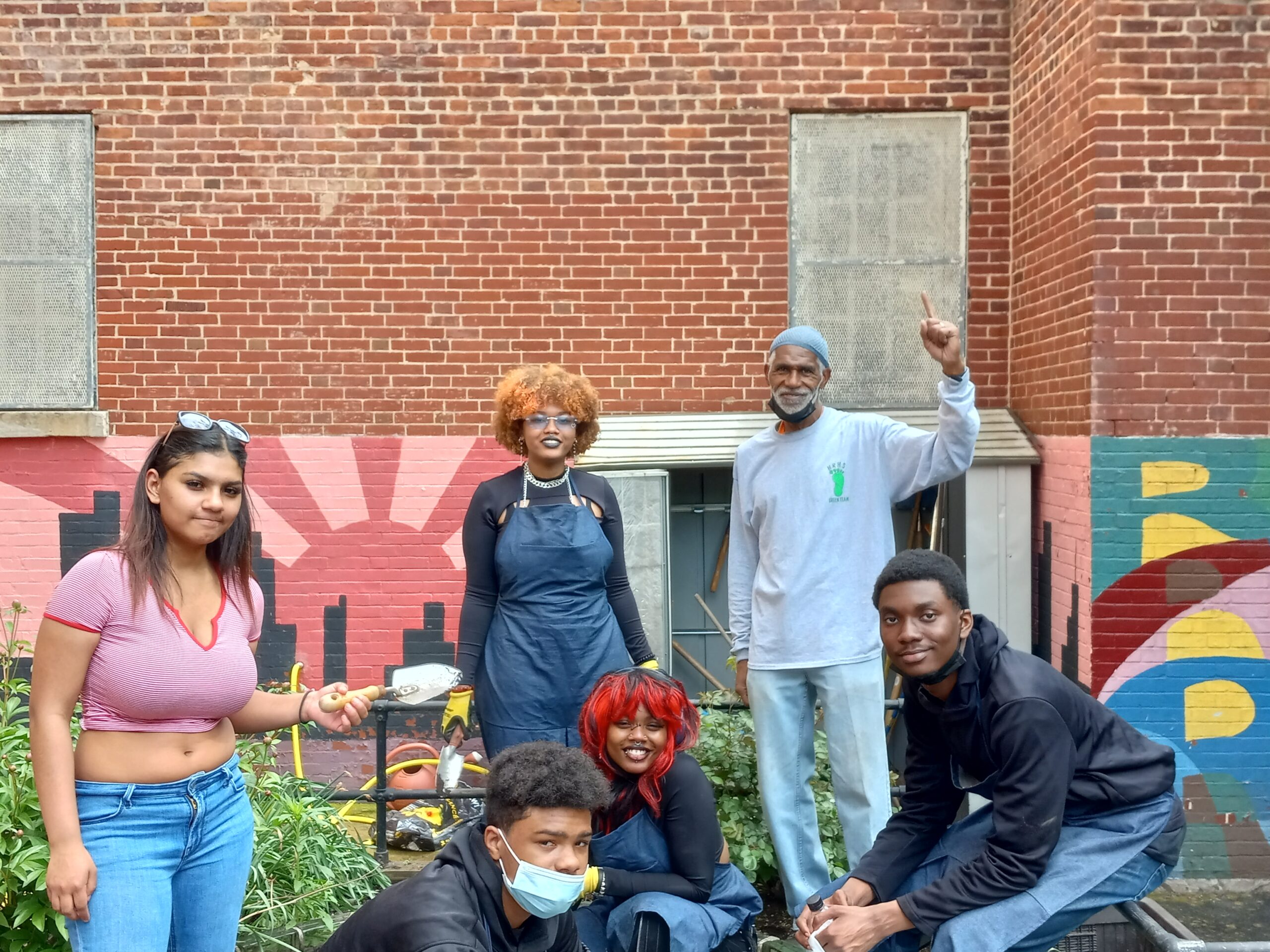Teacher Earl Grey and four high school students stand in front of a colorful mural on a brick wall posing with their freshly weeded school garden beds.